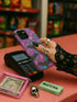 A protective phone case featuring a stained glass-style design in shades of purple, blue, and pink, with black bats flying across geometric panels outlined in gold. The case has a smooth matte finish and fits a triple-camera smartphone model.
